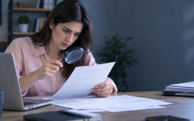 a person examining a document closely with a magnifying glass or a concerned, scrutinizing expression, papers and a laptop on a desk.